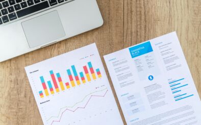 Top view of a laptop, charts, and resume on a wooden desk, showcasing business analysis and job application.